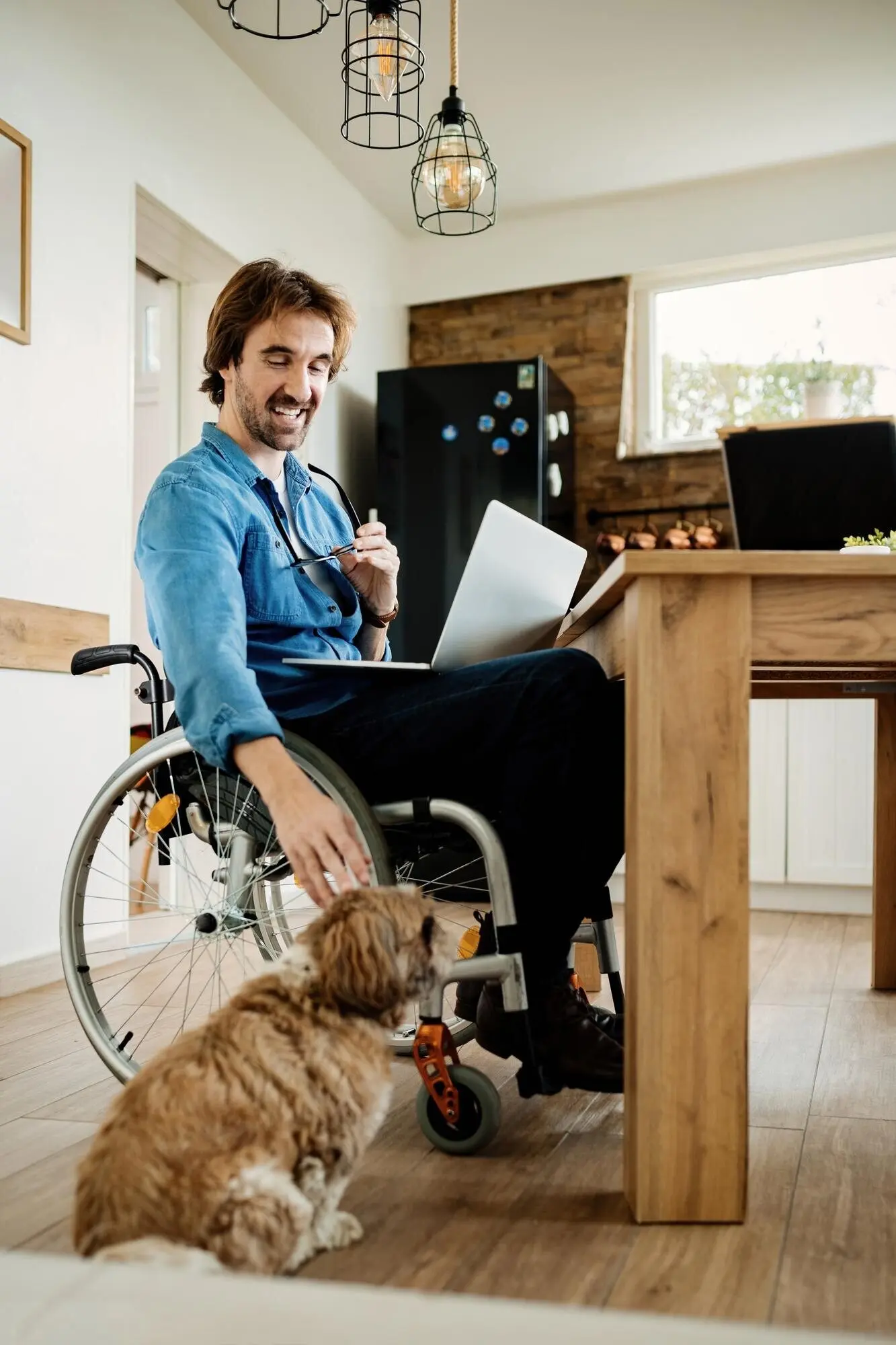 A happy young freelance worker in a wheelchair is working on a computer at home while cuddling his dog.