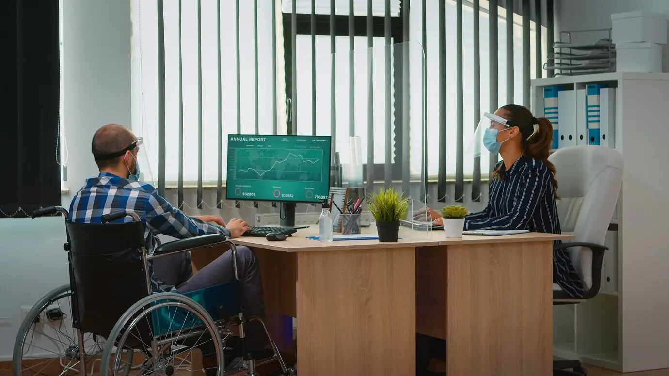 A manager with a disability arrives at the workplace in a wheelchair, wearing a protective mask, working in a new normal business office. An immobilized freelancer in a financial company respects social distancing.